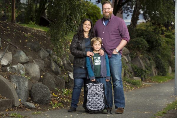 Family photograph, posed, mother and father stand looking at camera with child holding backpack in front of them. 