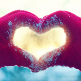 Red-gloved hands forming a heart shape in fresh snow, symbolizing holiday giving and community support.