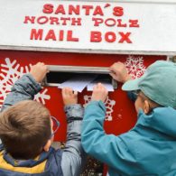 Two children place handwritten letters into a red “Santa’s North Pole Mail Box,” surrounded by holiday decorations.