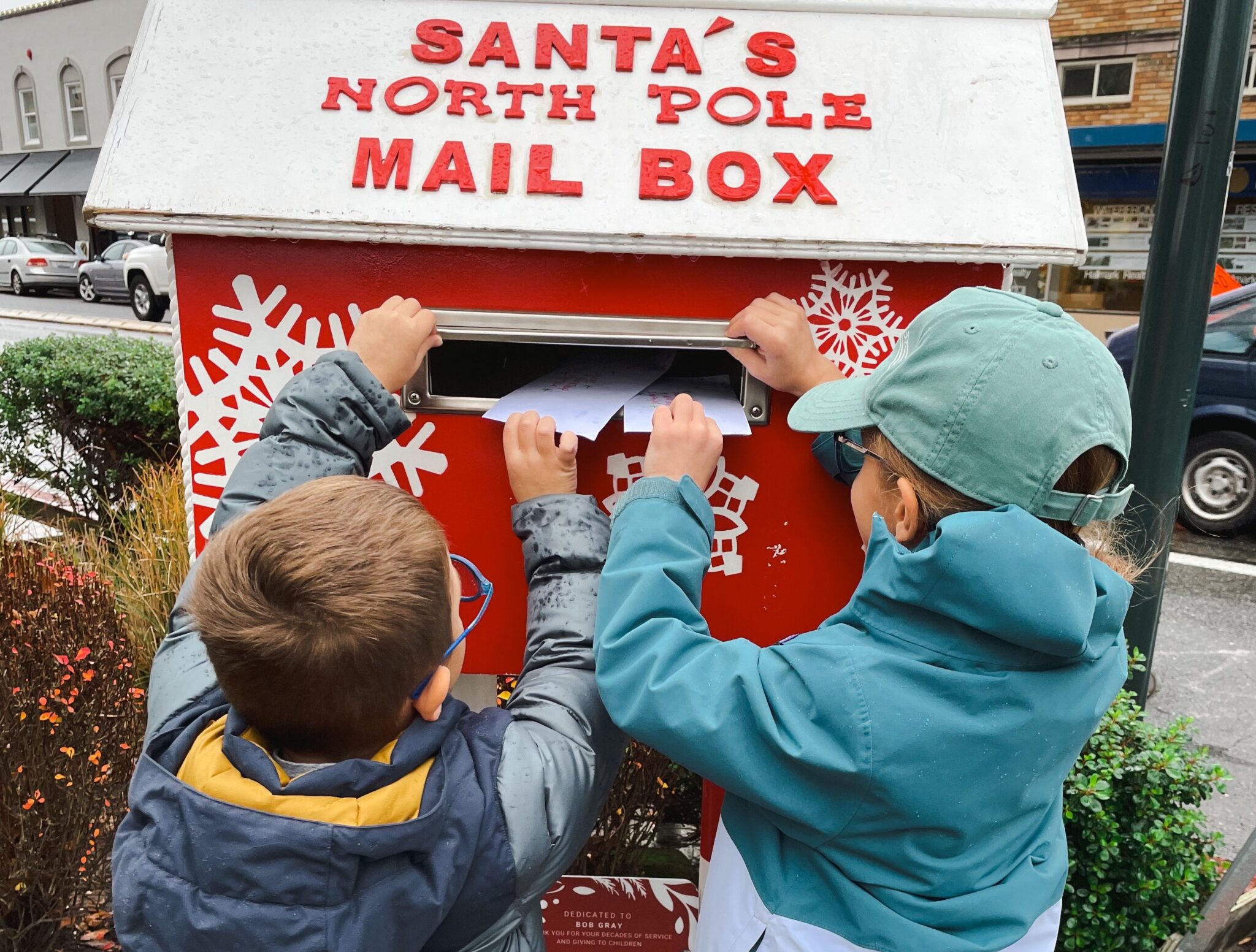 Two children place handwritten letters into a red “Santa’s North Pole Mail Box,” surrounded by holiday decorations.