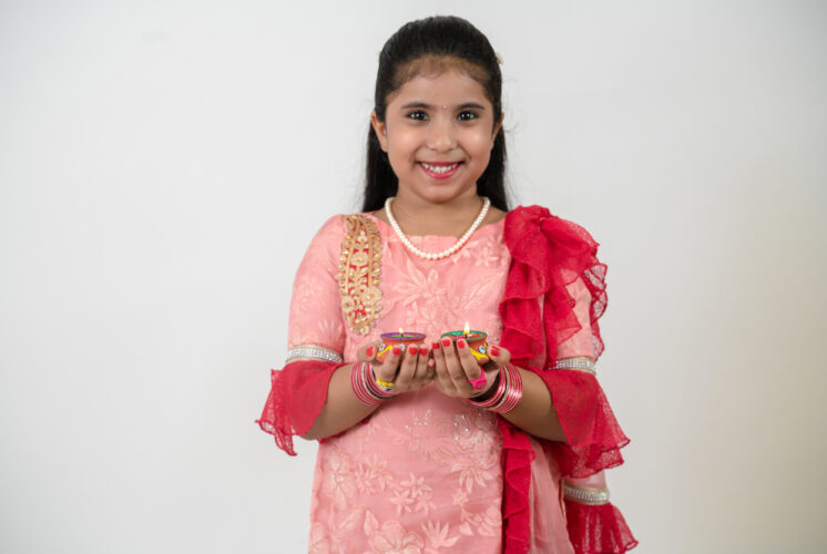 Young girl in traditional Indian dress proudly holding her colorful Diwali paper craft, surrounded by bright festive decorations.