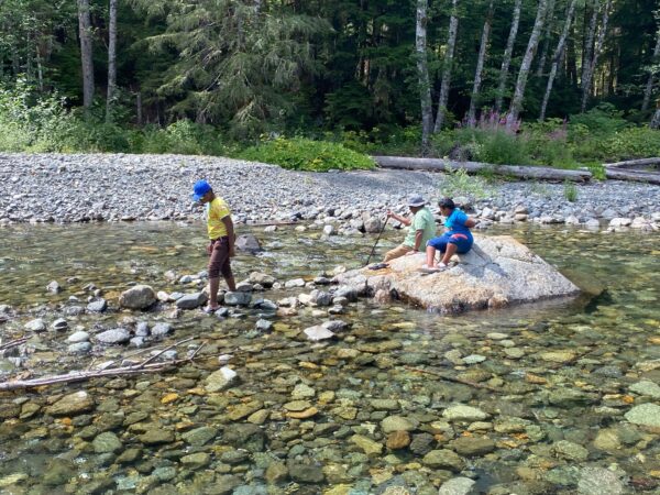 Children walking through and playing in the shallow Stillaguamish River near the Big Four Ice Caves trail, surrounded by rocks and water.