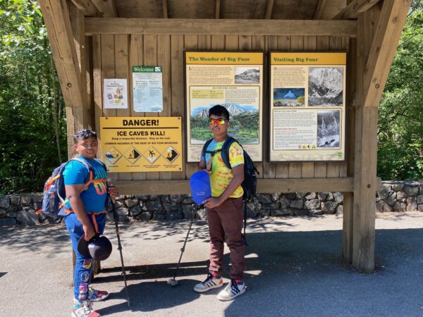 Two children standing in front of the Big Four Ice Caves trailhead sign, smiling and ready to begin their hike.