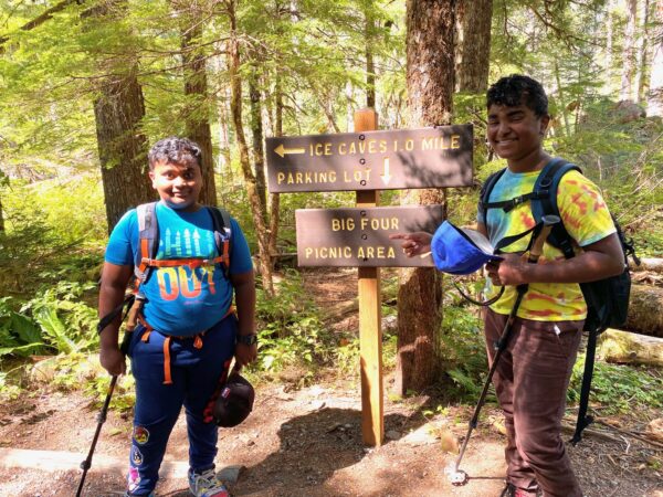 Two kids with hiking gear and poles standing next to a trail sign that reads “Ice Caves 1 mile, Parking Lot, Big Four Picnic Area.”