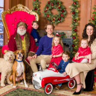 A family with four children and two dogs pose for a holiday photo with Santa, all smiling together in front of festive decorations.