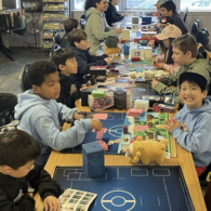 A group of children sit around a table at Tabletop Village in Seattle, playing Pokémon card games during a family game night.