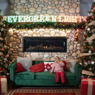 Girl sitting on a couch in front of a decorated fireplace with stockings and Christmas trees at Evergreen Christmas Lights in Bothell.
