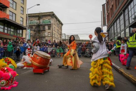 Two performers mid lion dance in front of a crowd outside on the street, wearing colorful Lunar New Year garb