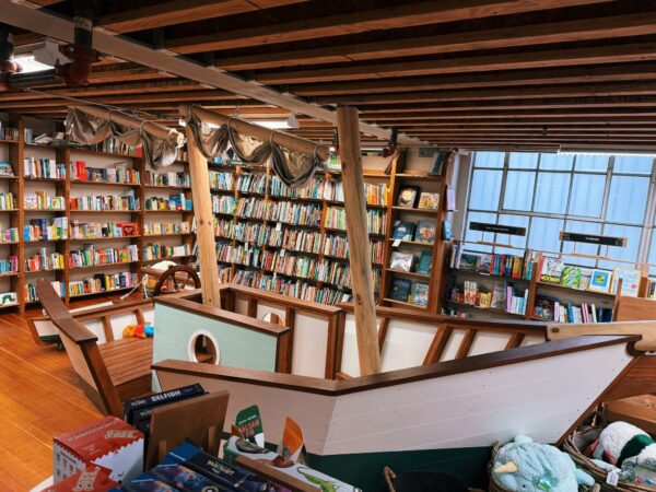 Large wooden play structure of a boat sits in the middle of a bookstore, surrounded by shelves.
