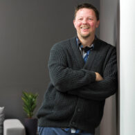 Man leans agains the wall of a sparse office, smiling at camera.