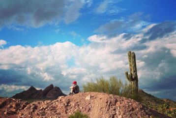 Boy with a red cap sits with his knees tucked in atop a desert hill, next to a cactus and under a bright blue sky.