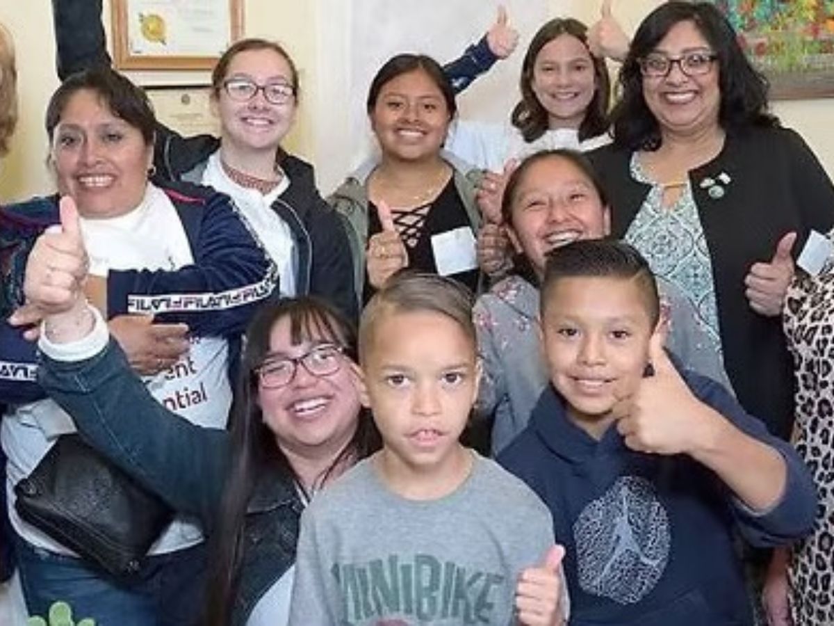 Group of kids and adults all huddle together facing the camera with thumbs up signs.