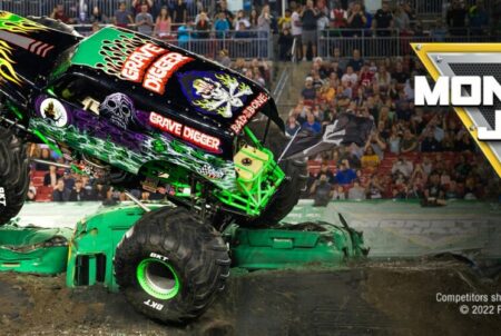 Huge green and black Monster Truck driving up the side of a dirt hill, in front of a crowd of people