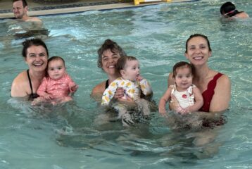 Three women holding babies in front of them, all facing the camera while standing in a pool.