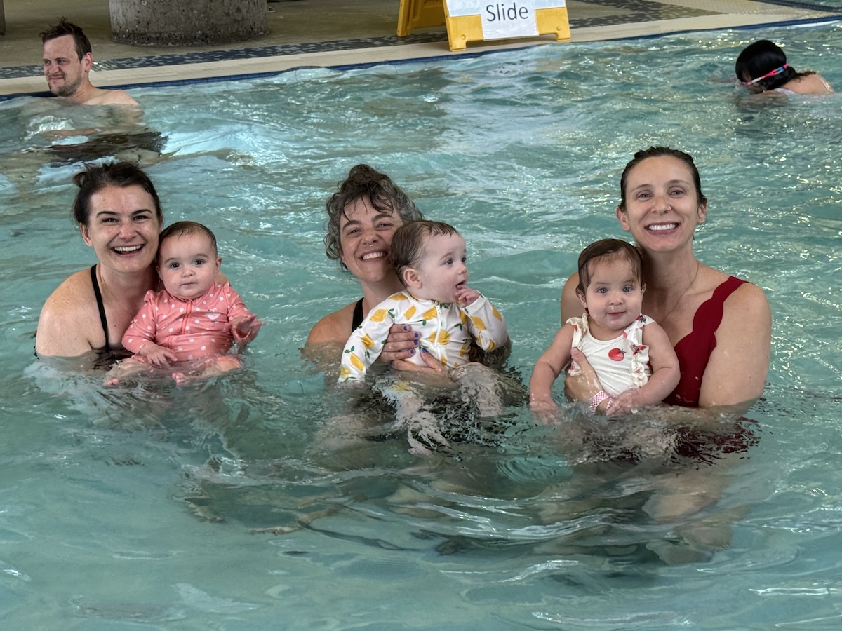 Three women holding babies in front of them, all facing the camera while standing in a pool.