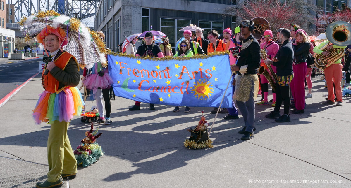 Brightly colored dressed people walk in parade with large Fremont Arts Council sign