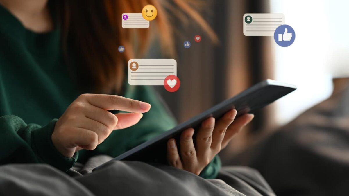 Close up of a hand touching a tablet, with Facebook likes and heart graphics surrounding the screen indicating social media use.