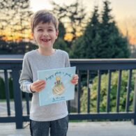 Boy, beaming, holds up a book titled Drippey Plants A Garden standing on his deck