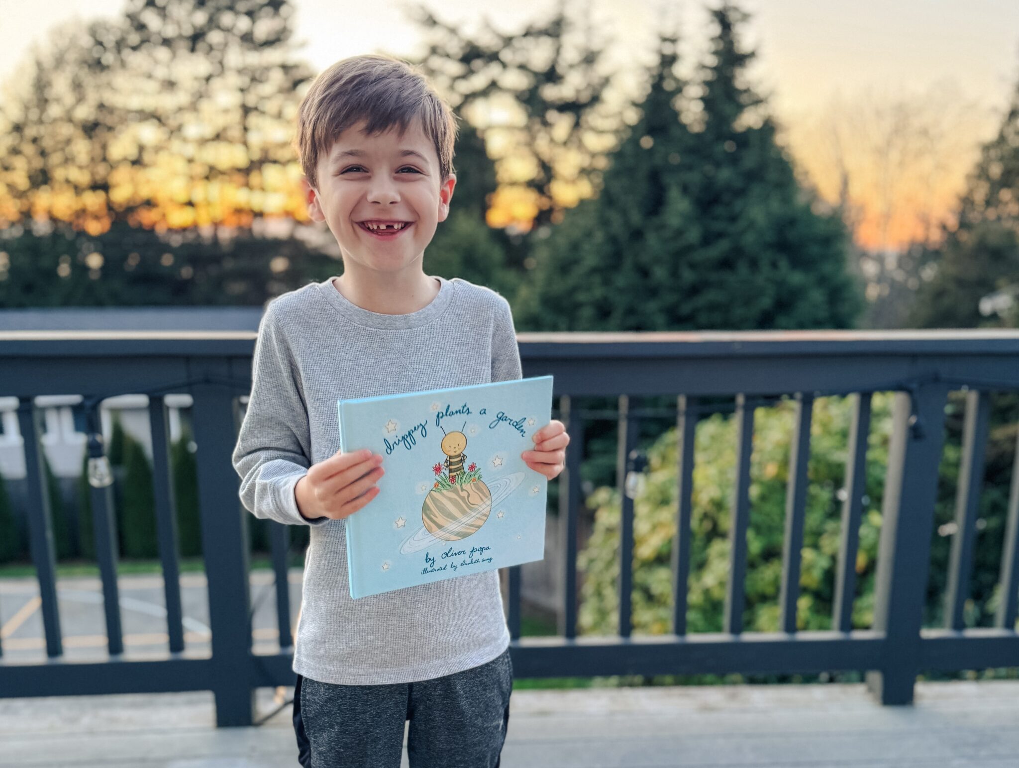 Boy, beaming, holds up a book titled Drippey Plants A Garden standing on his deck