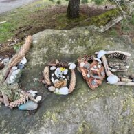The word 'LOVE' spelled out with driftwood, rocks, kelp, flowers, crabshells, pinecones. Sitting on a large boulder in the park