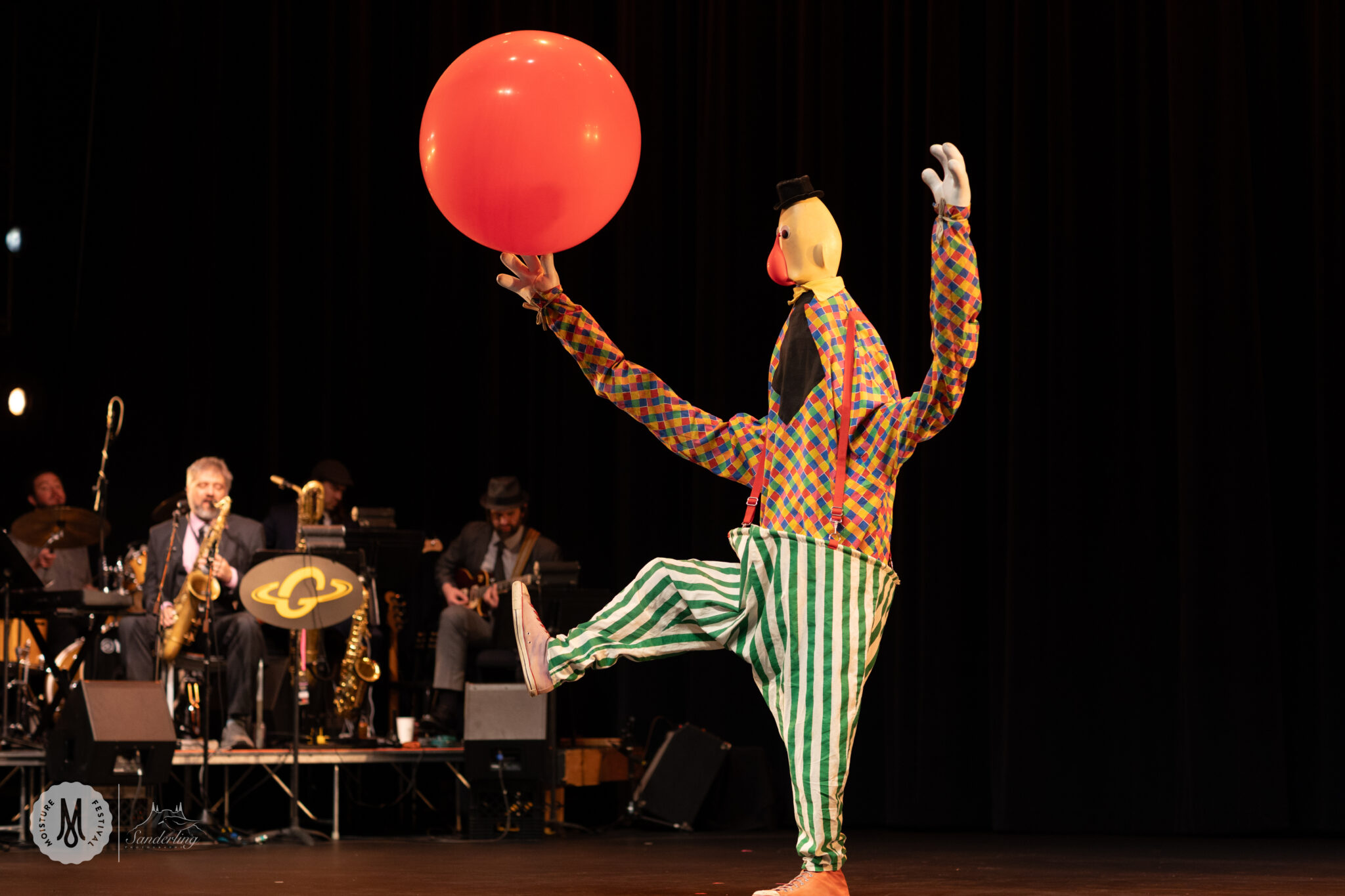 Costumed and masked performer wearing clownish costume hold big ball with foot up in kicking motion.