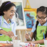 Asian and African American girls paint together during an art lesson. They are wearing aprons.