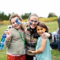 Two children and an adult share a warm hug at a Light Collective gathering, smiling with colorful face paint and surrounded by a joyful, welcoming atmosphere.