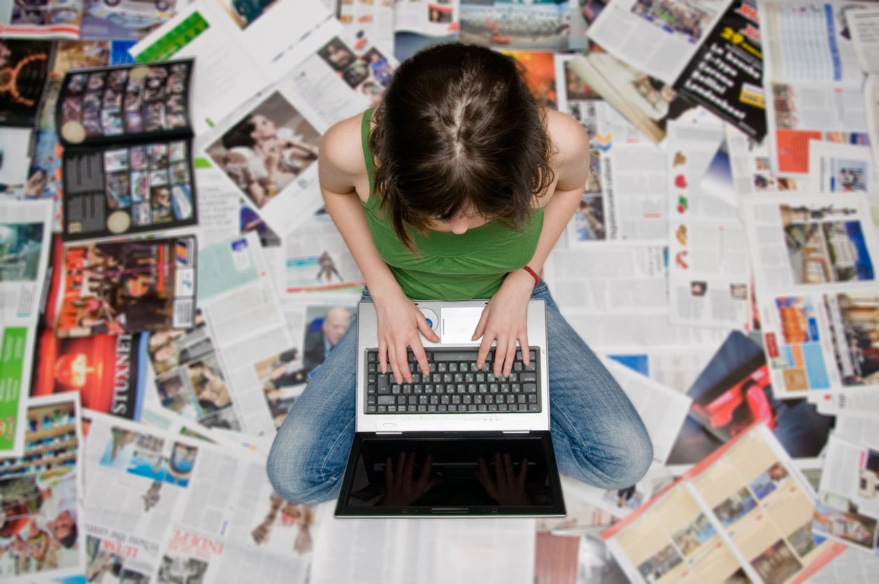 A student sits cross-legged on the floor surrounded by printed newspaper pages, typing on a laptop — symbolizing student journalism and creativity in Washington schools.