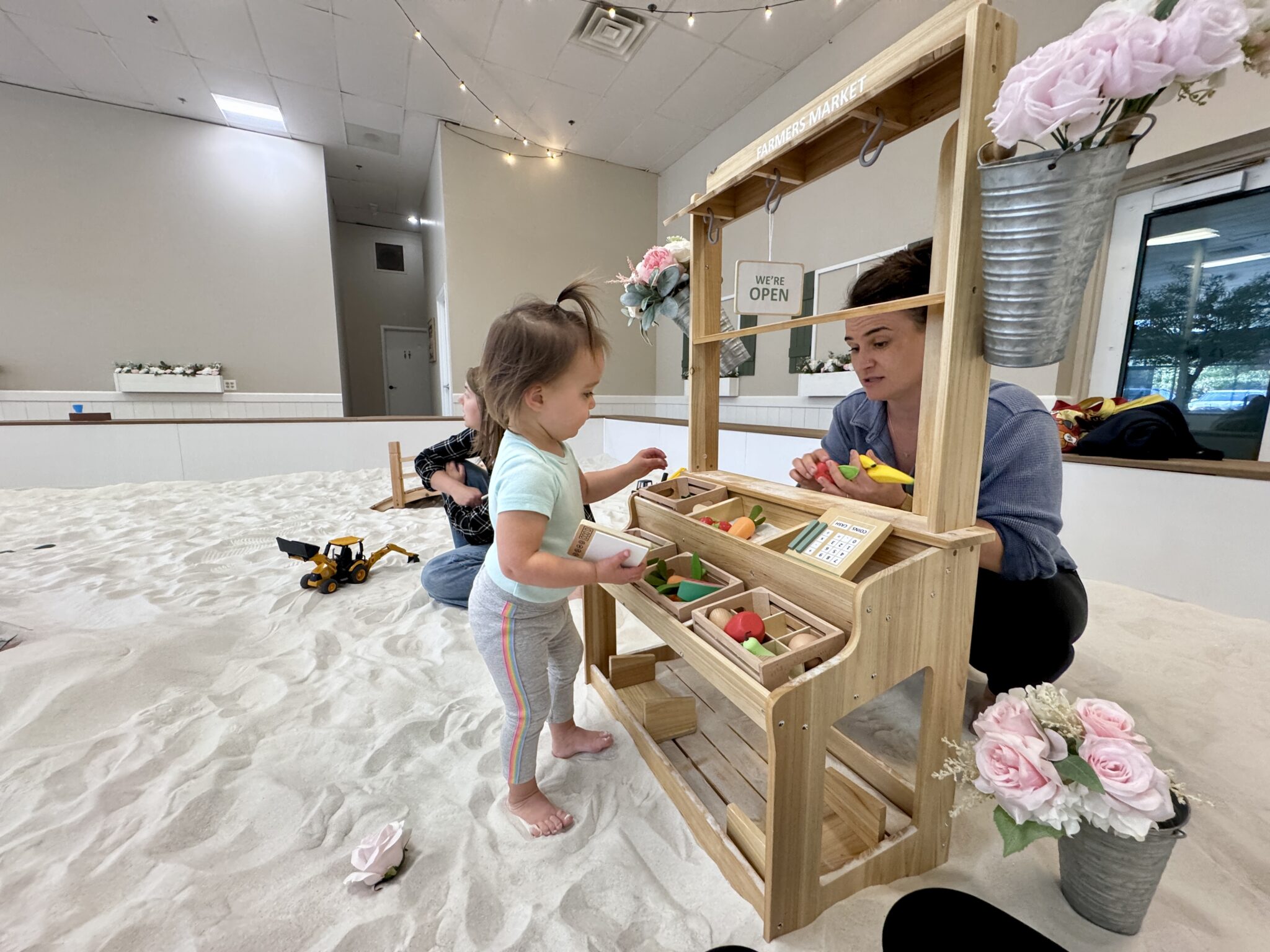 Toddler Ava and parent playing together with a toy grocery stand in Sand Garden’s sandbox