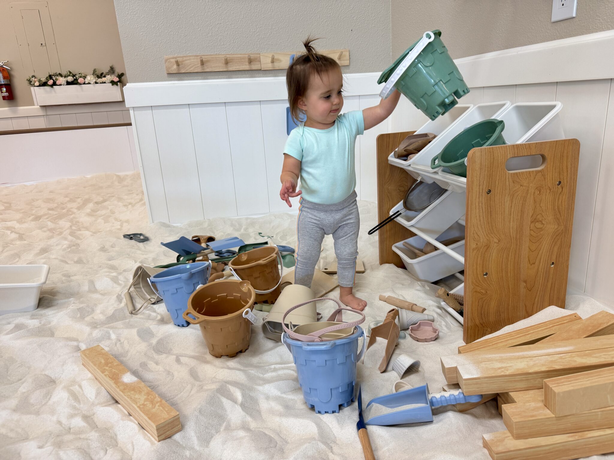 Toddler Ava pulling toys off a shelf in SandGarden’s sandbox area, with shovels, buckets, and cups scattered around her