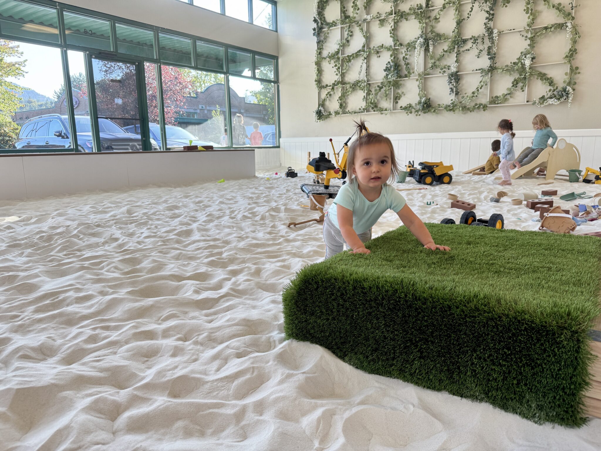 Toddler Ava standing barefoot in Sand Garden’s sandbox, looking up at the camera