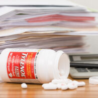 A tipped over and spilled bottle of Extra Strength Tylenol Acetaminophen Caplets in front of a pile of paperwork, a calculator, a pen, and the bottle lid. The Tylenol bottle contains 100 caplets, 500 mg each