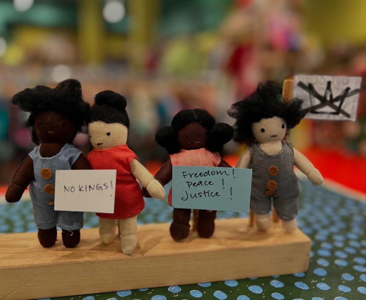 Four handmade cloth dolls stand side by side holding miniature protest signs reading “NO KINGS!” and “Freedom! Peace! Justice!” in front of a colorful background at Chrysanthemum, a children’s store in Seattle.