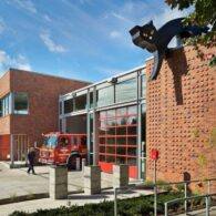 Exterior of Seattle’s Fire Station 9 with bright red bay doors and a large black-cat sculpture crouched on the rooftop, overlooking the sidewalk as a firefighter walks nearby.