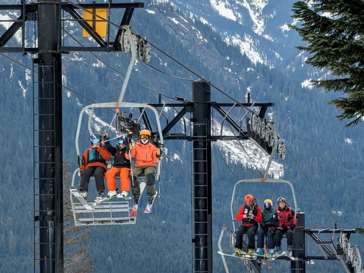 Families riding a chairlift at the Summit at Snoqualmie on a winter day, with skiers and snowboarders bundled in colorful gear and the snow-covered Cascade Mountains rising behind them.